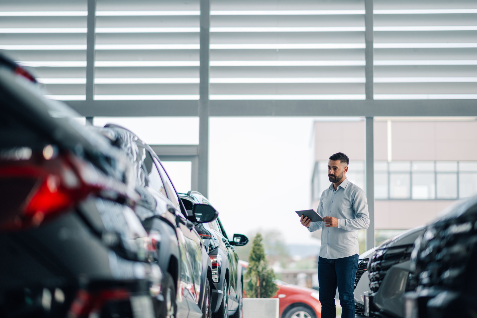 Man in grey shirt assessing cars with tablet in dealership view.